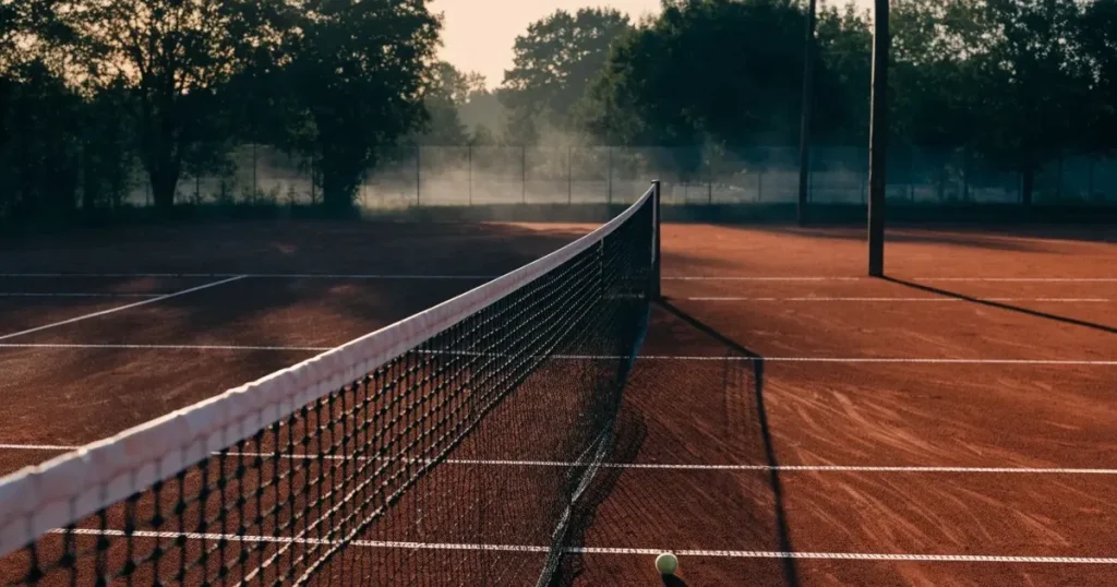 Ein Tennisplatz aus Sand bei Sonnenaufgang mit gespanntem Netz, Nebel zwischen den Bäumen am Rand