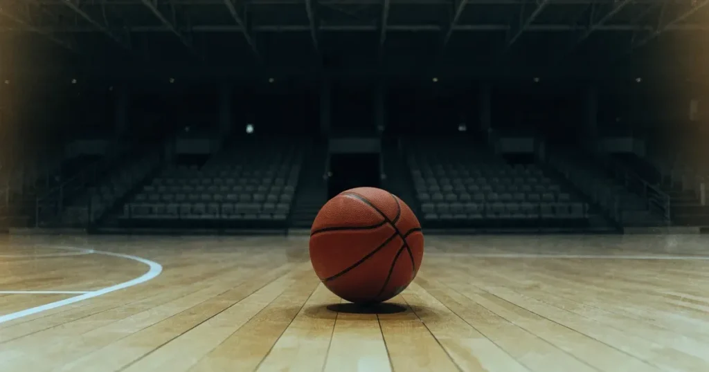 Ein Basketball liegt auf dem hellen Parkett einer leeren Halle, im Hintergrund steigen die Tribünenreihen im Halbdunkel an.