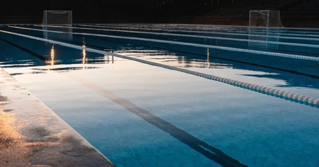 Ein Wasserballbecken am Abend mit zwei schwimmenden Torrahmen an den Enden, ruhiges blaues Wasser, leere Ränge.